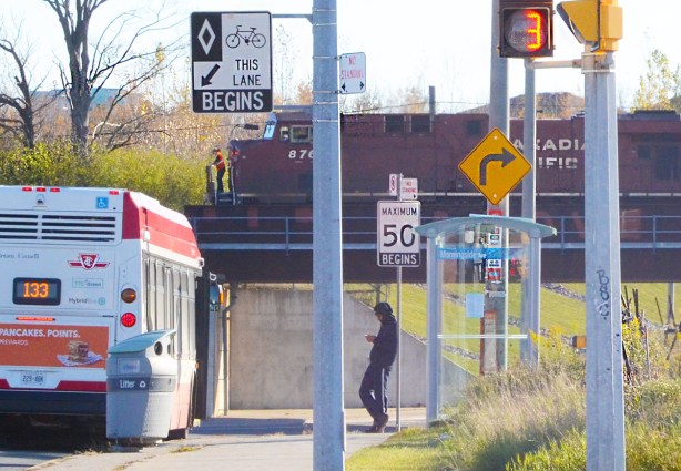 Canadian Pacific railway engines pass over bridge over Finch Ave. A TTC bus is waiting in the foreground, as well as a man standing at the bbus shelter. 