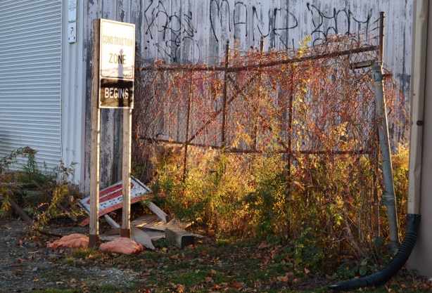 construction ahead sign off to the side beside a chainlink fence with weeds growing behind it, afternoon sun is shining through fence