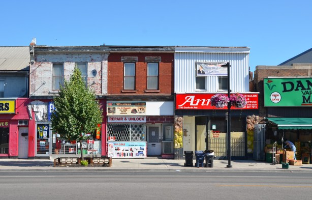 a row of three two storey brick stores on the Danforth.  Flat roofs, one in the middle is a computer repair shop.  A small tree covers the front of one store, Danforth market is on the far right