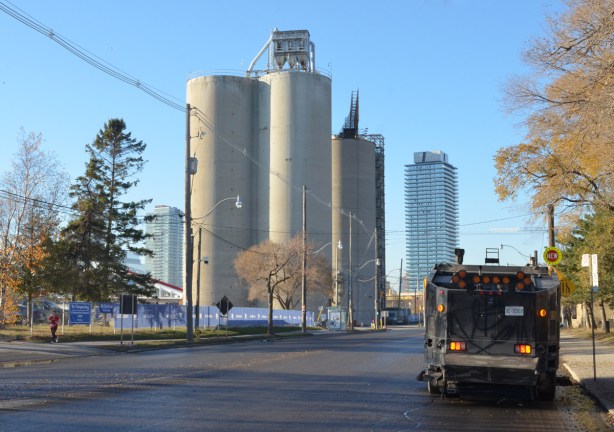 three tall silos that were part of essroc cement plant, now a heritage site in the port lands, a street cleaner is parked on the road, two condo towers in the distillery district can be seen in the background 