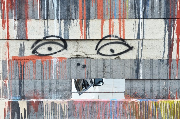 dripping paint in blue and red on a wall with a hole in it, two eyes drawn above the rectangular hole. A car is parked on the other side of hte wall and shows through the hole 