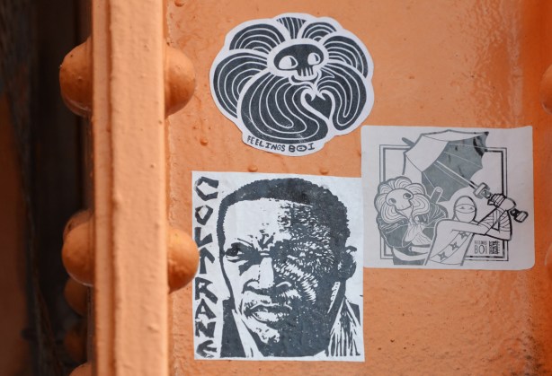 three stickers on orange metal support of west toronto railpath bridge over Bloor. One is a portrait of a black man Coltrane, another is a feelings boi sticker and last is a sticker that is feelings boi along with urban ninja squadron, 