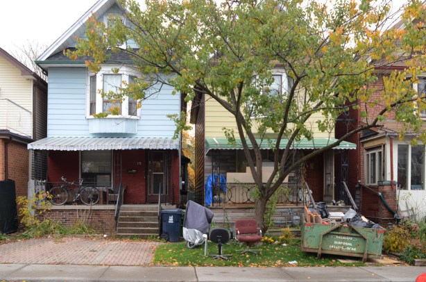 2 houses side by side, one with pale blue siding on upper floor, the other with yellow siding, both with porches in the front, the one n the right is being renovated and has a bin out the front 
