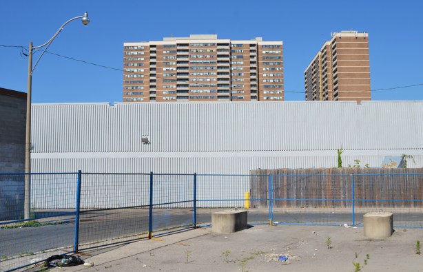 tall apartment buildings behind a tall plain grey wall, a street in front and then a vacant lot that's been paved over 