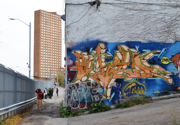 highrise apartment building in the background with a concrete building with lower level covered by street art in the foreground, people walking in the alley behind the concrete building 