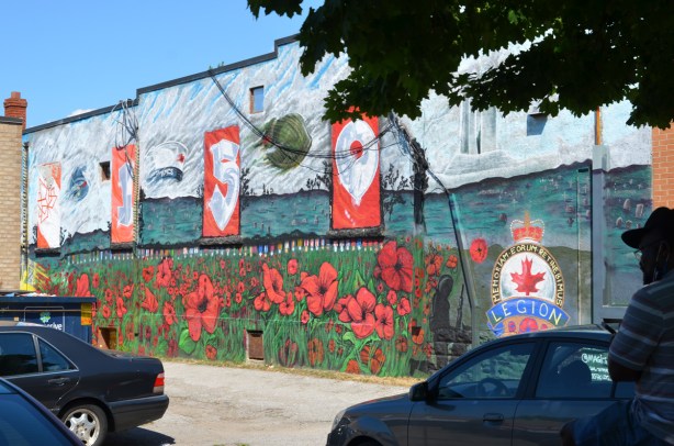 cars parked and a man sitting in front of mural in the side of Royal Canadian Lion branch 73, Oakridge Branch, poppies, a yellow airplane, beside an alley, 