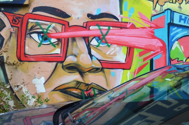 part of a uber5000 mural of a person wearing red glasses, also reflected in the windshield of the car that is parked beside the wall 