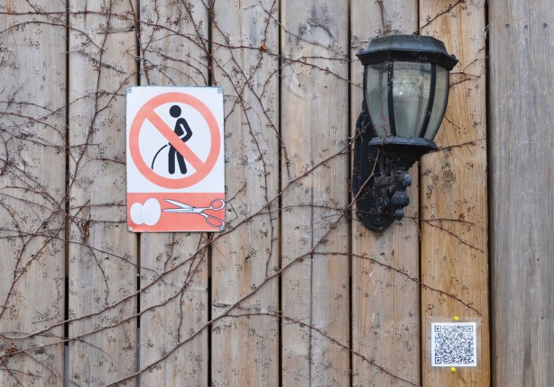 wood fence, exterior light, and a sign, picture of man peeing with a red line through it, below that is picture of a pair of scissors and two eggs. 
