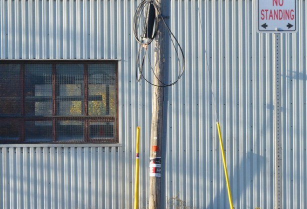 a grey utility pole and a small pole with a no standing sign in front of a light grey metal building with a window in it 