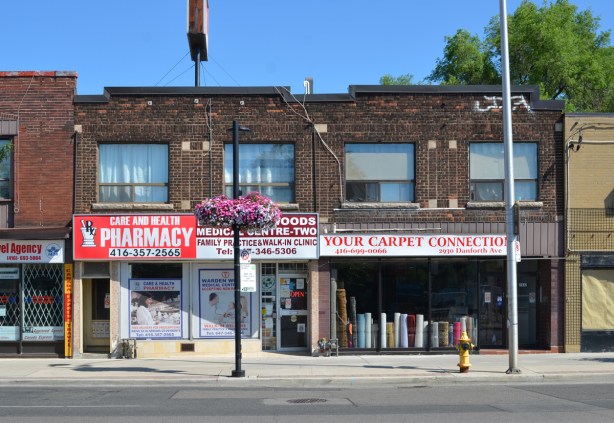 old two storey brick building on Danforth, stores, pharmacy, walk in clinic, and carpet store, your carpet connection with rolls of carpet in the window