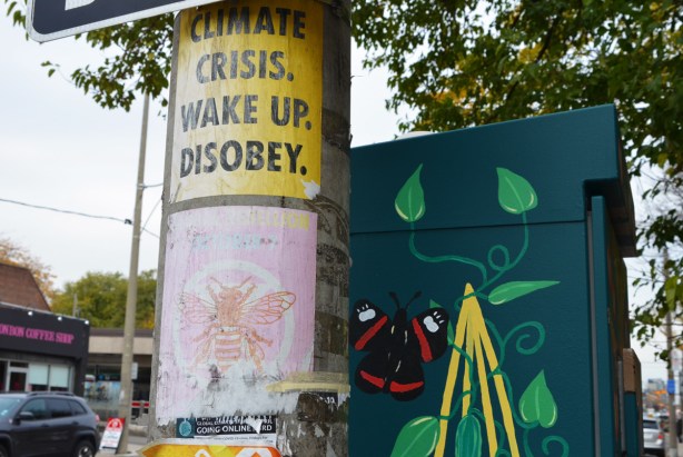 2 posters on a utility pole plus a painted street box behind. One poster says climate crisis wake up disobey. The other poster has a pink bee