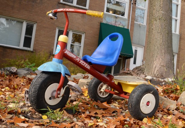 a radio flyer tricycle parked on the grass, with lots of fallen leaves, in front of brick apartment building 
