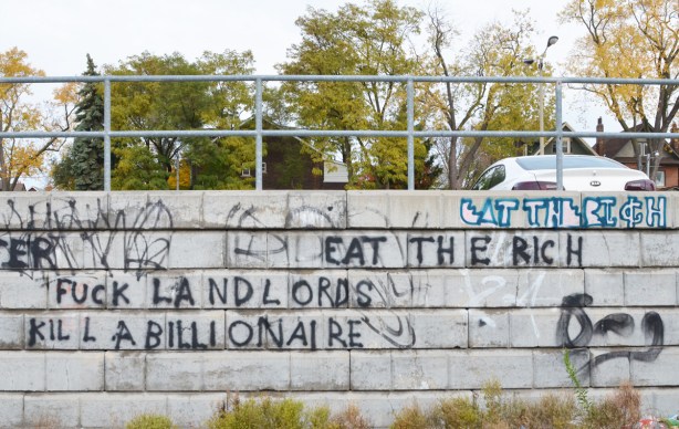 words scrawled on a concrete wall by a parking lot. eat the rich, kill a billionaire, fuck landlords, 