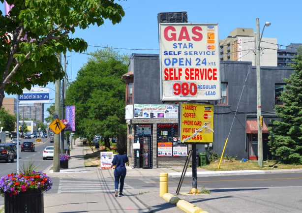 woman walking on Danforth at Madeleine, past star self service gas station, large sign with price of gas as 98 cents a litre, computer store on opposite side of street 