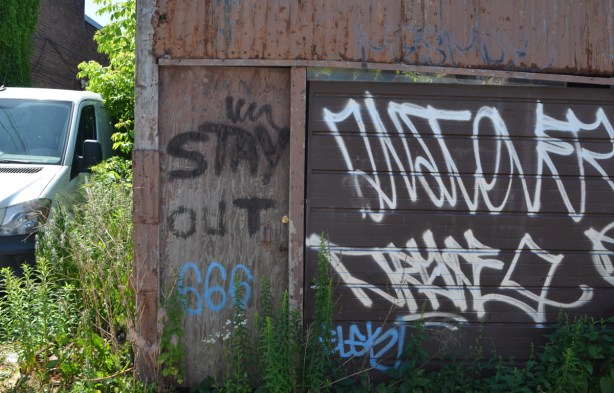 old wood door beside metal garage door, outside, in an alley, large words spray painted on door that say stay out 666
