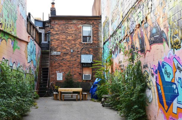 back of two storey brick building with a small paved space behind it, a table and benches are near the building, street art on both sides of the small space (the buildings on either sode) 