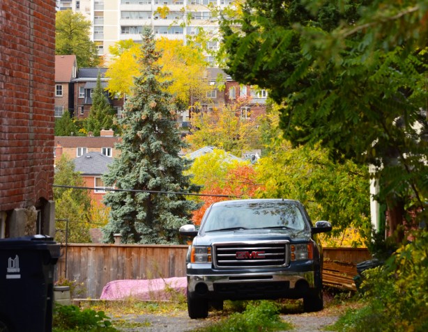 a G M C truck is parked in a gravel driveway between two buildings, in the background is a couple of rows of houses and a tall apartment building behind that 