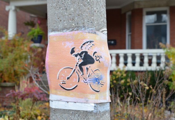 stencil on paper on a utility pole, in orange and black, a girl riding a bike, with wings on her back 