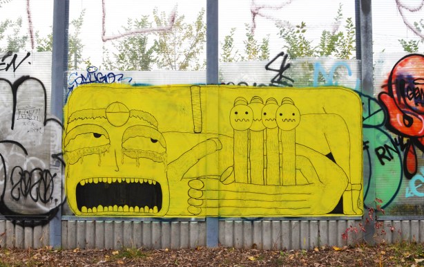 yellow street art painted on glass wall beside West Toronto Railpath and railway tracks, large face with mouth open, hand holding 4 little figures that look like fingers 