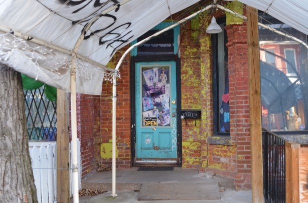 turquoise door with window it. window is covered with pictures, entrance to store is small sidewalk that is covered with a temporary portable tent like structure, 