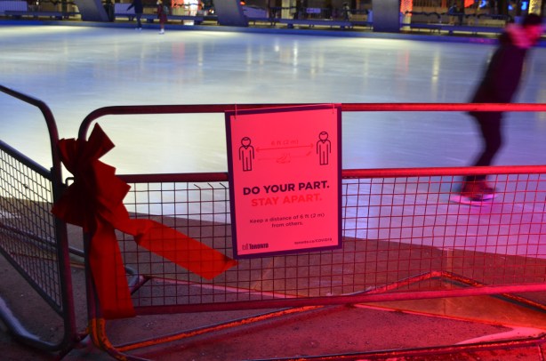 red flood lit metal barricade beside rink at Nathan Phllips Square, with a big red bow on it as well as a sign that says do your part, stay 2 metres apart, covid sign