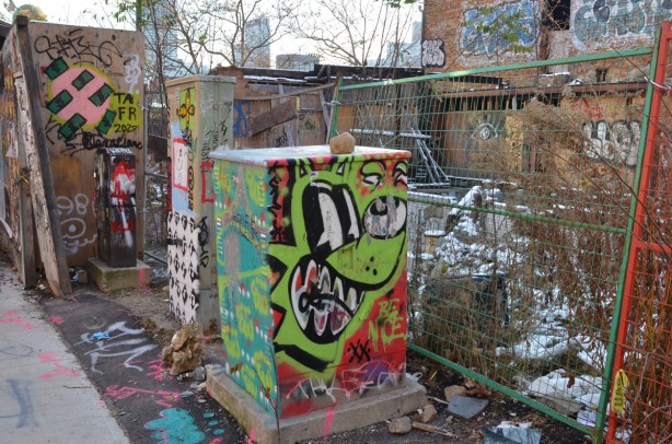 street art on metal boxes on sidewalk in front of a vacant lot with partially destroyed building on it 