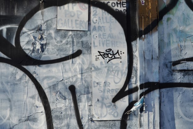 graffiti and posters on a wall that were painted over in silver gray paint but you can see traces through the paint. including a sign for a pop up store 