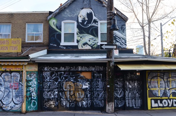 building covered with street art, two storeys, windows above, store below. Greys. panels covering store windows are all covered with painting too 