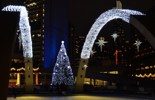 christmas lights and stars on the arches over the skating rink at Nathan Phillips square, with the large Christmas tree covered in blue and white lights behind. Also, part of the towers of city hall are lit blue