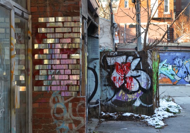 mirrors in the shapes of bricks glued on top of some bricks on an exterior wall in an alley, also a fence with graffiti 