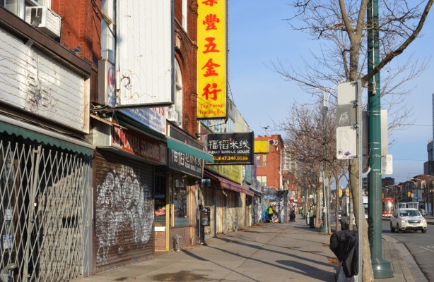 storefronts on Spadina ave., a large yellow sign with red chinese characters for Tap Phong trading company, empty sign, storefront with metal gate covering entrance, 