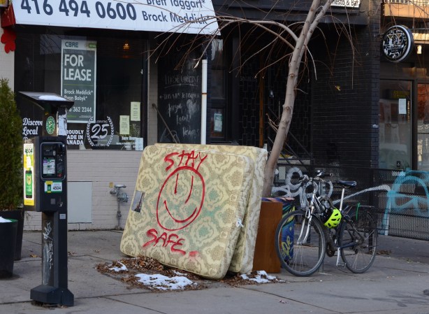 2 old mattresses left on the sidewalk, leaning against a small tree. big happy face spray painted onto one of them along with the message stay safe 