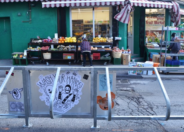 across the street, people are getting a fruit and vegetable store ready to open up, putting food on display outside. in the foreground is a metal bike stand with graffiti slaps on it, including a urban ninja squadron and soap ghost, wash your hands 