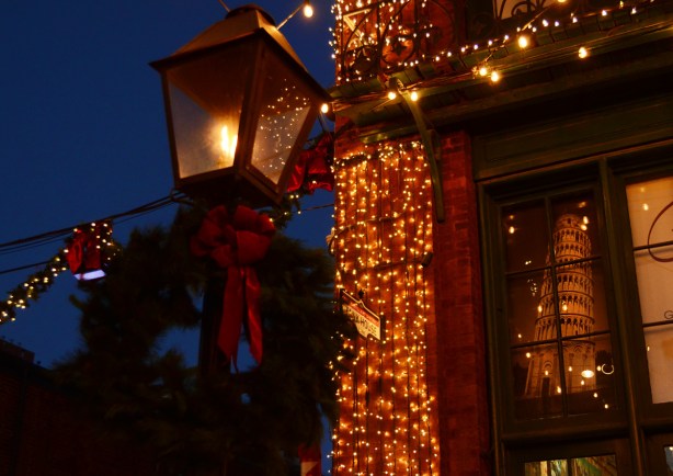 evening photo, distillery district, decorated for Christmas, a street lamp that looks like an old gas lamp, with a wreath and a big red bow, beside a mens wear store with a picture of the Leaning Tower of Pisa in it 