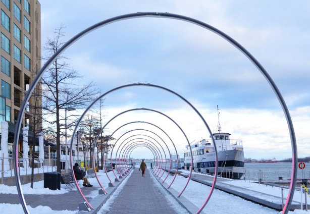 rings with a pinkish colour surround a walkway, a woman is walking through them, on the waterfront, a boat is docked beside the walkway