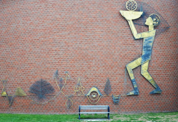 metal 2 dimensional sculpture by Lionel Thomas mounted on a red brick wall, title is The Whole Person, a man is holding a burning lamp