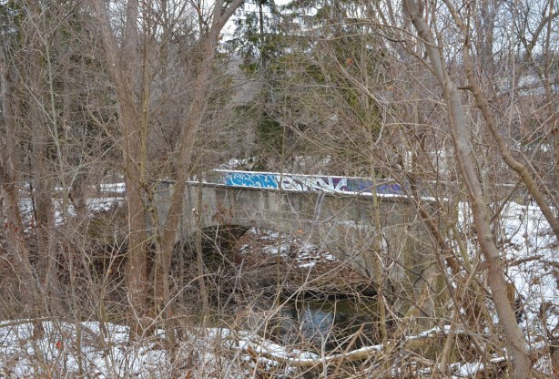 an old concrete bridge over the Don River by Bayview, some graffiti on it, seen through the woods in winter, no leaves on the trees, over the Don River, 