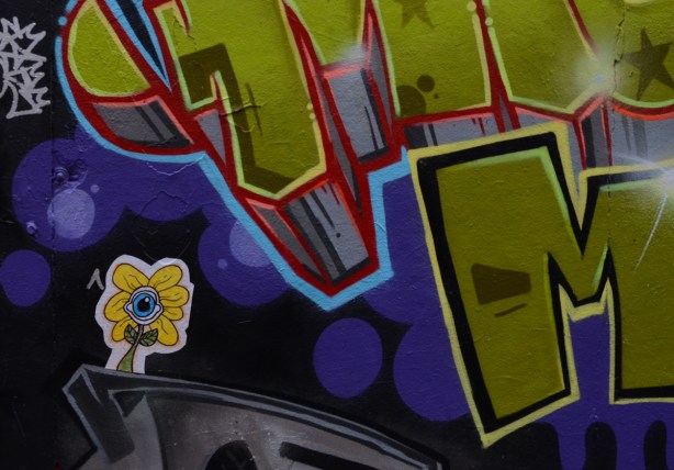 small sticker of a daisy with yellow petals and an eye in the center, on a wall already covered with painted street art 