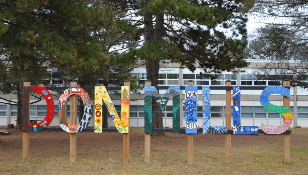 Painted sign tht says Don Mills, in front of Don Mills Sceondary school. Each letter is a different colour with decorations on them (pictures) painted by students 