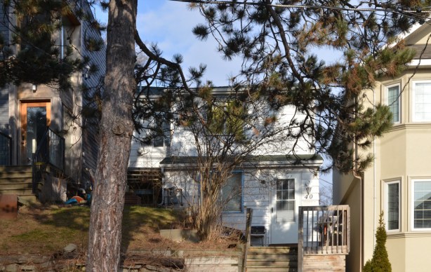 small white house behind a large pine tree and between two larger houses, on a small hill with stairs leading up to it 