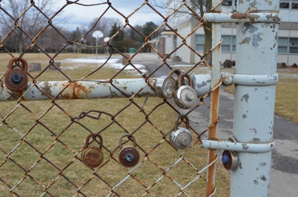 a few locks, rusted, on the chainlink fence around Don Mills Secondary school, playing fields and basketball hoops in the background 