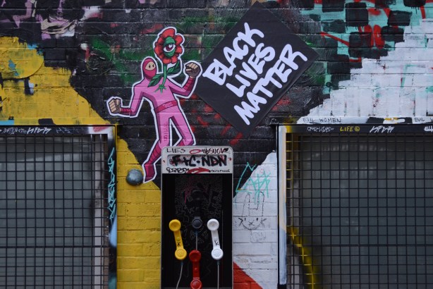 black lives matter words on a wall with an urban ninja squadron t bonez in a pink suit and holding a red daisy with one eye in the center, graffiti alley