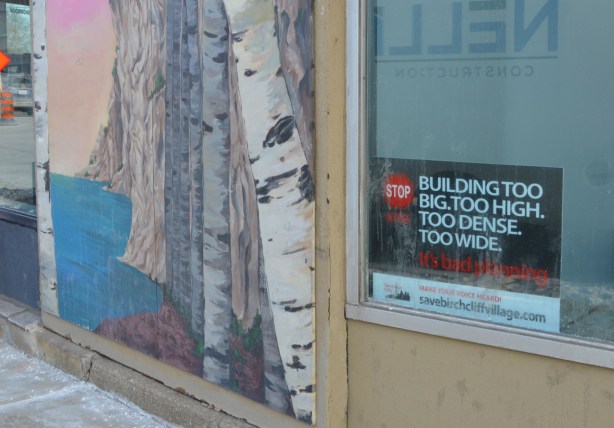 small mural of birch trees beside Scarborough bluffs, on outside wall beside a window with a protest sign in it. Poretesting redevelopment of parts of Kingston Road with buildings that are too big, too tall, too wide 