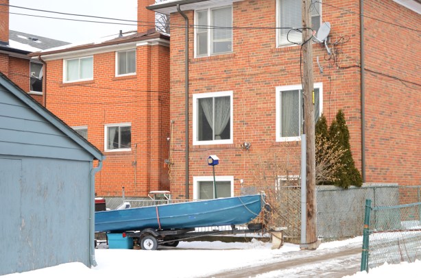 small blue boat on a trailer parked by garage in an alley behind multiplex houses 3 storeys high, red brick. 