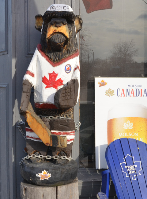 carved wooden bear, Buster, with Canada flag hockey shirt on, holding a hockey stick, 