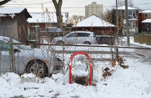 a chair in the driveway by an alley, in the snow, cars, chainlink fence behind the chair. 