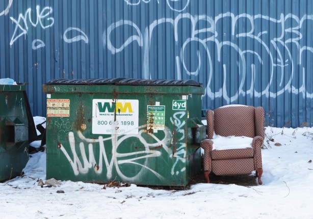 an old beige arm chair, with snow it, outside beside industrial garbage bins 