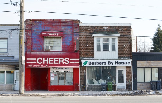 three old two storey brick storefronts, one is Cheers restaurant painted bright red, the other is Barbers by Nature