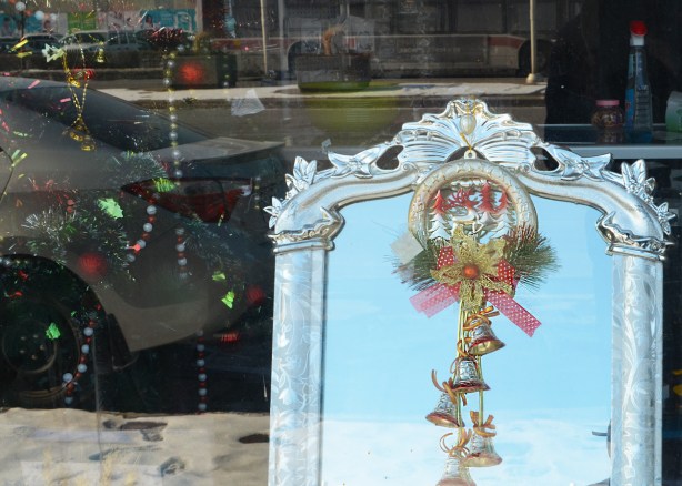 looking in the window of a store, a mirror with an ornate silver colour frame, Christmas bells attached to it with ribbons and greenery