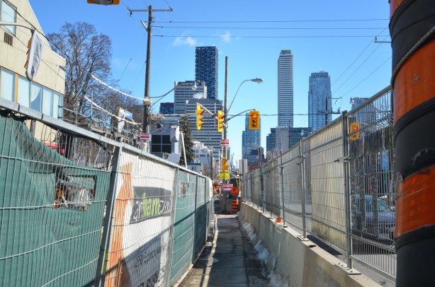 Eglinton Ave west sidewalk through Crosstown construction, barriers on both sides, narrow, tall buildings at Yonge and Eglinton in the background
